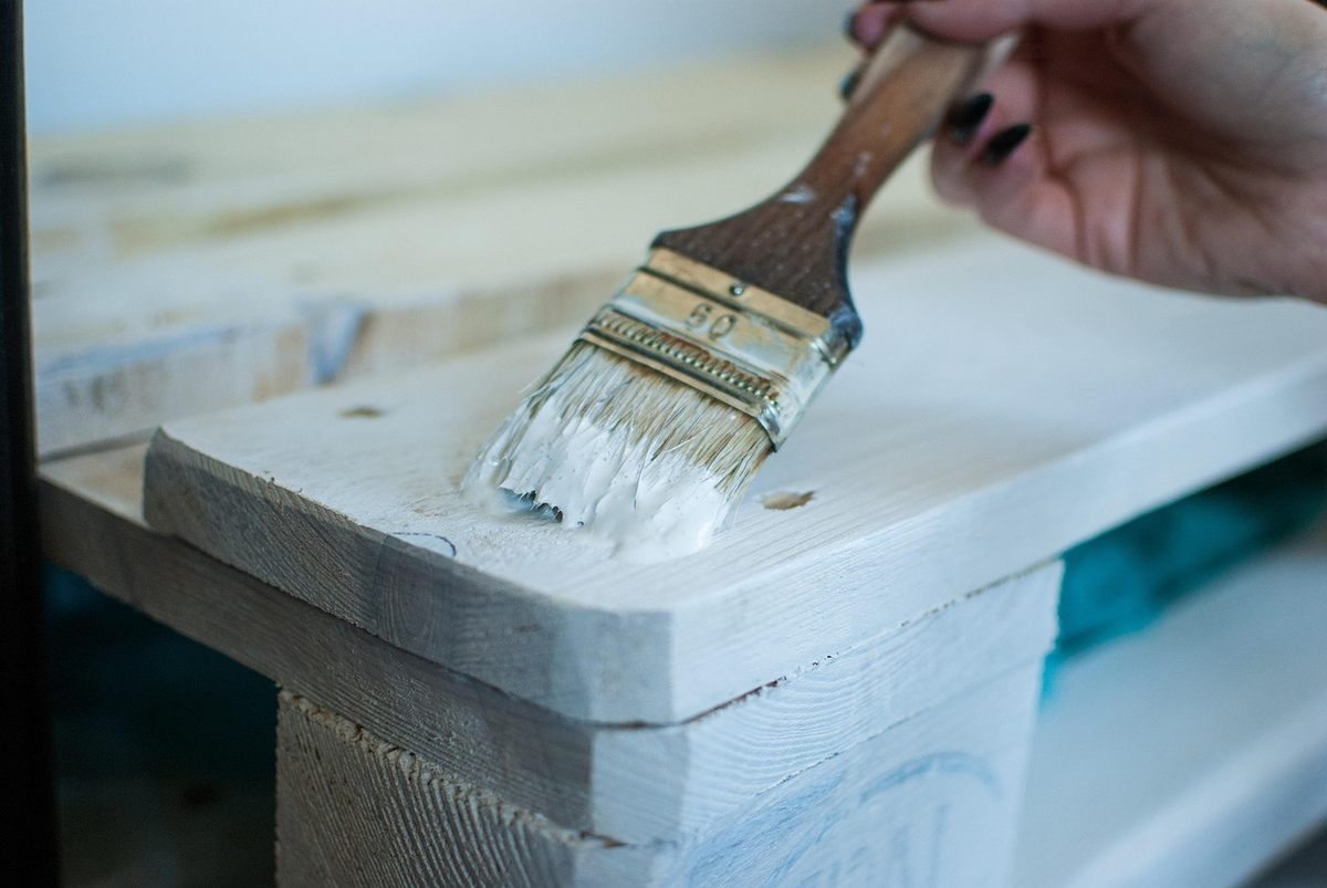 Main tenant un pinceau appliquant de la peinture blanche sur une surface en bois stratifiée Main tenant un pinceau appliquant de la peinture blanche sur une surface en bois stratifiée